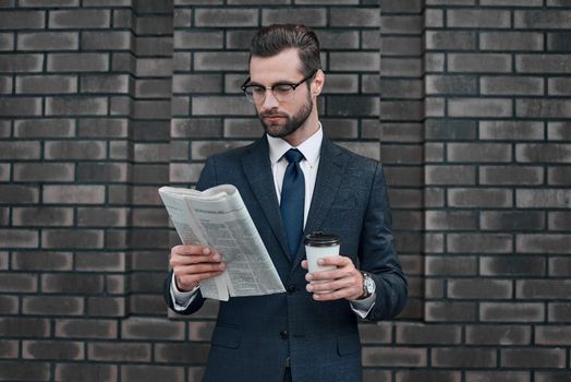 Checking business news. Good looking young man in full suit reading a newspaper on the wall background