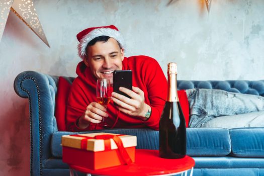 Young handsome man in Santa Claus hat with glass of champagne conducting live broadcast with friends on smartphone. Happy guy celebrating Christmas online on internet and lying on couch at home