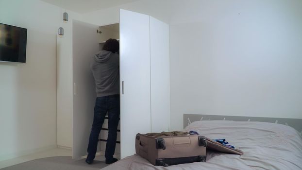 Young man using smartphone talking with friend and packing suitcase at home. Guy wearing in casual hoodie pack up clothes in travel or business trip. Male walking near wardrobe in flat.