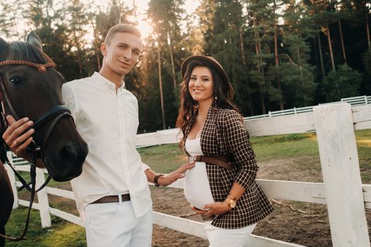 a pregnant girl in a hat and a man in white clothes stand next to horses near a white fence.Stylish pregnant woman with a man with horses.Married couple