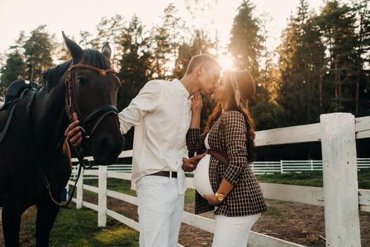 a pregnant girl in a hat and a man in white clothes stand next to horses near a white fence.Stylish pregnant woman with a man with horses.Married couple