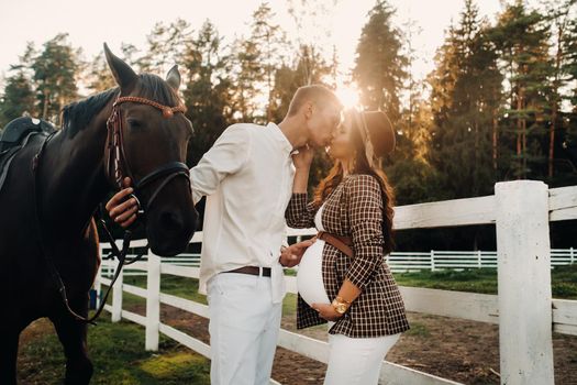 a pregnant girl in a hat and a man in white clothes stand next to horses at a White fence.Stylish family waiting for a child strolling in nature.