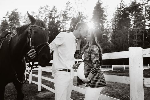 a pregnant girl in a hat and her husband in white clothes stand next to the horses near the horse corral at sunset.Stylish pregnant woman with a man with horses.Family. black and white photo.