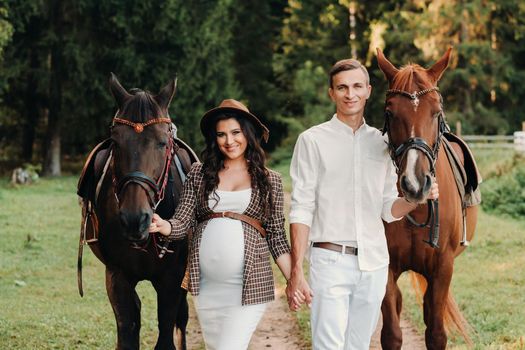 a pregnant girl in a hat and her husband in white clothes stand next to horses in the forest in nature.Stylish pregnant woman with a man with horses.Family