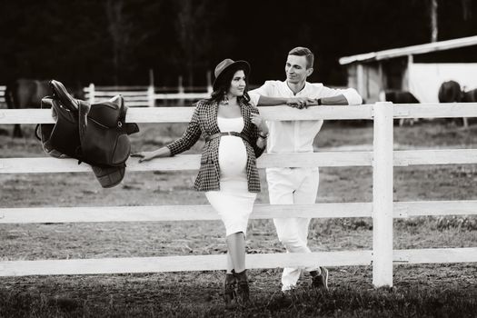 a pregnant girl in a hat and her husband in white clothes stand next to the horse corral.a stylish couple waiting for a child stand on the street near the horse corral. black and white photo.