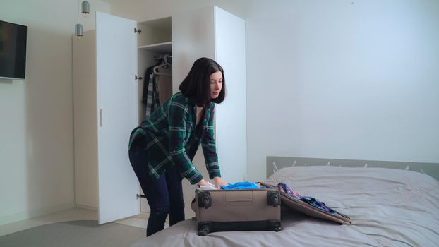 Young woman packing suitcase at home. Happy female pack up clothes in travel or business trip. Brunette choose what she want dress in vacation.