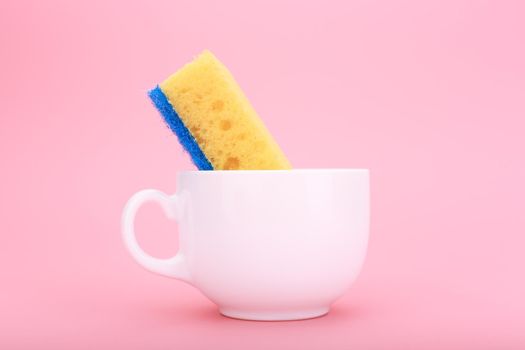 Creative, minimal dishwashing concept. Simple composition with yellow cleaning kitchen sponge in white ceramic cup on pink background 