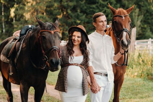 a pregnant girl in a hat and her husband in white clothes stand next to horses in the forest in nature.Stylish pregnant woman with a man with horses.Family