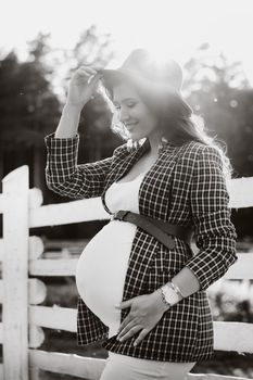 a pregnant girl with a big belly in a hat near a horse corral in nature at sunset.Stylish pregnant woman in a brown dress with horses.black and white photo.