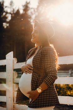 a pregnant girl with a big belly in a hat near a horse corral in nature at sunset.Stylish pregnant woman in a brown dress with horses