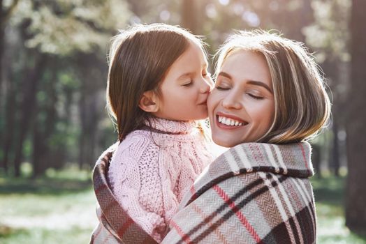 A little curly girl in sweater is kissing her mother in woods. She is wearing pink sweater and young woman is in grey sweater. They are wrapped up with a blanket