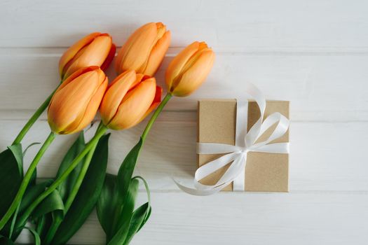 A beautiful bouquet of orange tulips and a small craft ingess with a white satin ribbon tied to a bow. Festive composition of spring flowers and gift box.