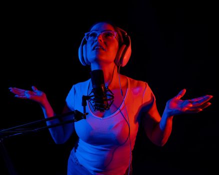 Caucasian woman in glasses and headphones sings into a microphone in neon light on a black background. An emotional girl is recording a song in a recording studio.