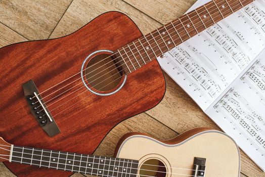 Music education. Brown acoustic guitar and ukulele lying on the wooden floor with music notes. Top view. Music equipment. Music shop. Musical instruments