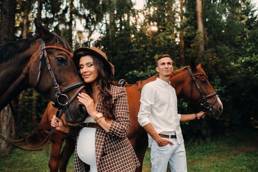 a pregnant girl in a hat and her husband in white clothes stand next to horses in the forest in nature.Stylish pregnant woman with a man with horses.Family