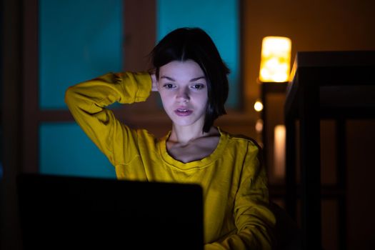 Portrait of beautiful caucasian girl uses laptop while sitting at home. In the evening creative woman works on a computer in her cozy living room.
