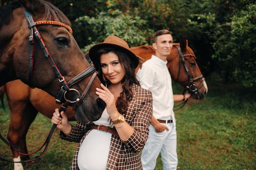 a pregnant girl in a hat and her husband in white clothes stand next to horses in the forest in nature.Stylish pregnant woman with a man with horses.Family