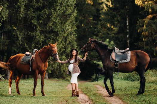 pregnant girl with a big belly in a hat next to horses in the forest in nature.Stylish pregnant woman in the brown dress with the horses