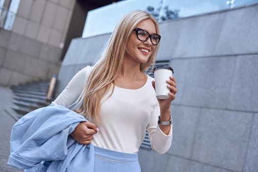 Happy young spectacled business woman is walking on the street with coffe. She is smiling. She is in stylish blue suit