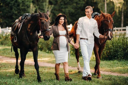 A pregnant woman in a hat with a man in white clothes walking with horses in nature. A family waiting for a child walks in the woods