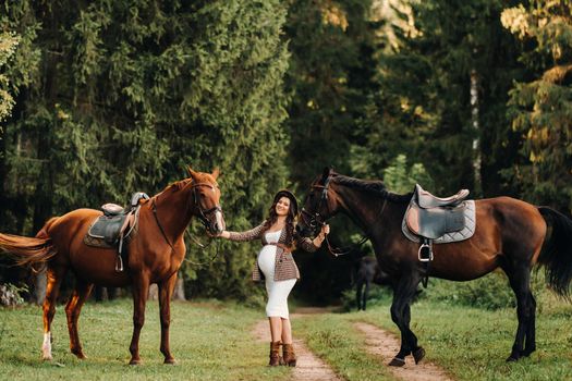 pregnant girl with a big belly in a hat next to horses in the forest in nature.Stylish girl in white clothes and a brown jacket