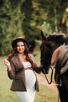 pregnant girl with a big belly in a hat next to horses in the forest in nature.A stylish pregnant woman in a white dress and brown jacket with horses