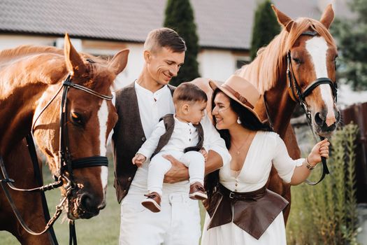 A family in white clothes with their son stand near two beautiful horses in nature. A stylish couple with a child are photographed with horses.