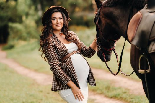 pregnant girl with a big belly in a hat next to horses in the forest in nature.A stylish pregnant woman in a white dress and brown jacket with horses