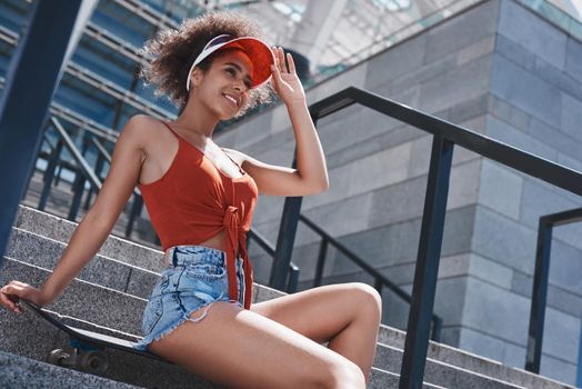 Young woman wearing tennis visor free style on the street sitting on concrete stairs on skateboard looking aside smiling happy relaxed