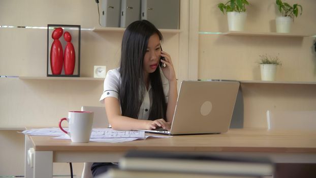 Asian designer talking on smartphone with client in the office. check information on computer.