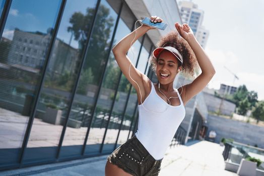 Young woman and earphones free style on the street walking near window holding smartphone listening to music dancing active closed eyes smiling joyful