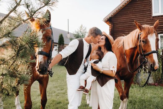 A family in white clothes with their son stand near two beautiful horses in nature. A stylish couple with a child are photographed with horses.