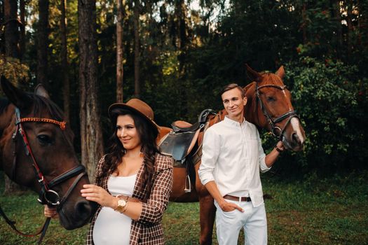 a pregnant girl in a hat and a man in white clothes stand next to horses in the forest in nature.Stylish pregnant woman with her husband with horses.Married couple