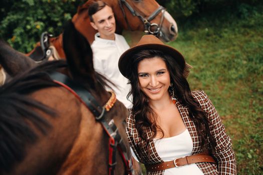 a pregnant girl in a hat and a man in white clothes stand next to horses in the forest in nature.Stylish pregnant woman with her husband with horses.Married couple