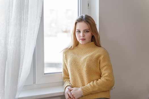 portrait of a beautiful teenage girl in a yellow sweater standing by the window