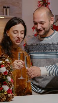 Festive couple talking on video call conference while holding glasses of champagne for christmas eve festivity. People using online communication and celebrating holiday with seasonal decorations