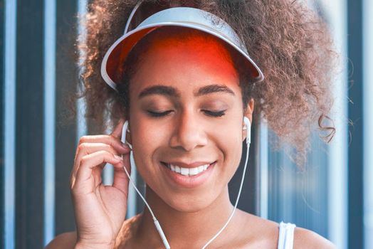 Young woman wearing tennis visor putting on earphones free style on the street standing near fence closed eyes smiling happy close-up