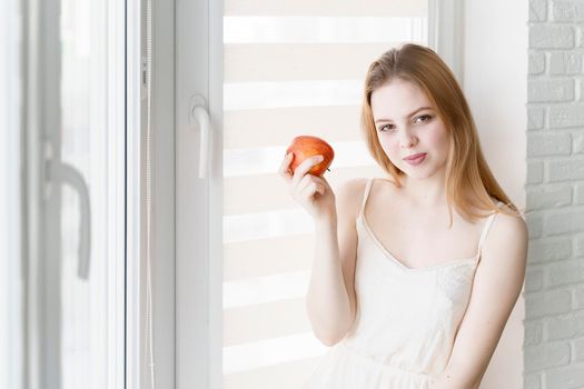 beautiful young woman in a white dress holding a red apple by the window in a bright room