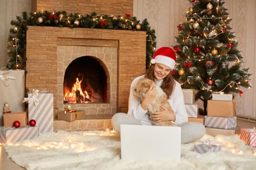 Happy girl in red hat having video call on laptop and sitting with cute dog at christmas tree with lights and presents in festive room, looking smiling ar device screen.