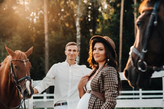 a pregnant girl in a hat and her husband in white clothes stand next to the horses near the horse corral.Stylish pregnant woman with a man with horses.Family.