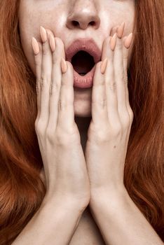 Studio shot of young and shocked redhead woman keeping mouth opened while standing against grey background. Surprise