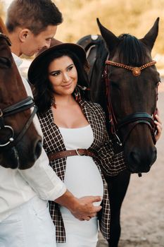 a pregnant girl in a hat and a man in white clothes stand next to horses near a white fence.Stylish pregnant woman with a man with horses.Married couple