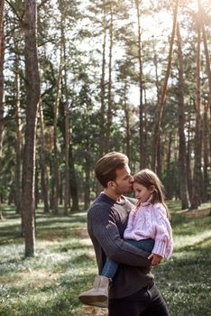 Little girl and her father in the forest. Young father kisses his little pretty daughter. Young man is wearing a dark sweater, girl is in pink bright sweater