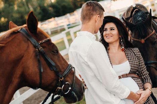 a pregnant girl in a hat and a man in white clothes stand next to horses at a White fence.Stylish family waiting for a child strolling in nature