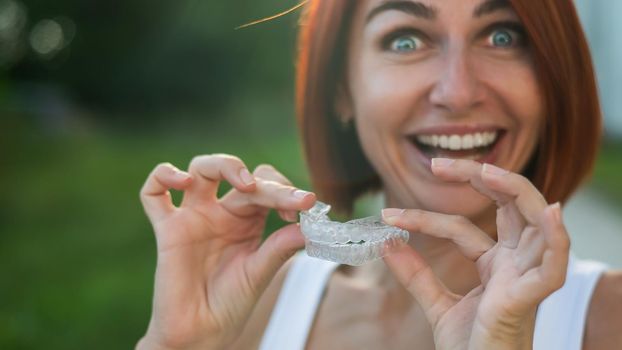 Red-haired Caucasian woman holding transparent mouthguards for bite correction outdoors. A girl with a beautiful snow-white smile uses silicone braces.