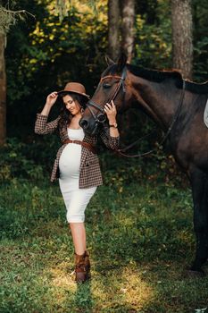 pregnant girl with a big belly in a hat next to horses in the forest in nature.Stylish girl in white clothes and a brown jacket