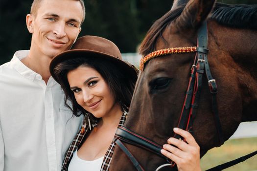 a pregnant girl in a hat and a man in white clothes stand next to horses at a White fence.Stylish family waiting for a child strolling in nature