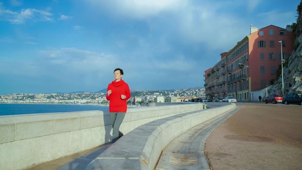 Female running by sea shore. Wearing red sports sweatshirt grey tights. Jogger listening music on earphones. Sunny day in spring season. On the background mediterranean city view.