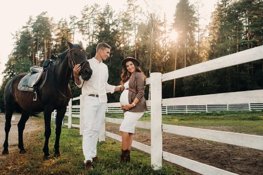 a pregnant girl in a hat and a man in white clothes stand next to horses at a White fence.Stylish family waiting for a child strolling in nature.