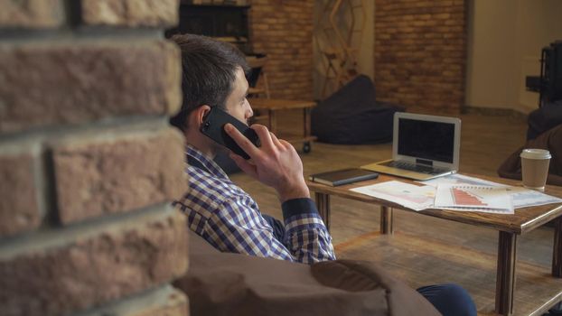 Analyst man talking on the phone on the table is a laptop, cup of coffee is, spread out the working papers. Soft panorama